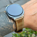 A close-up of a smartwatch with a round silver-toned case and a tan leather strap worn on a person's wrist. The background shows blurred outdoor greenery and gray pavement.