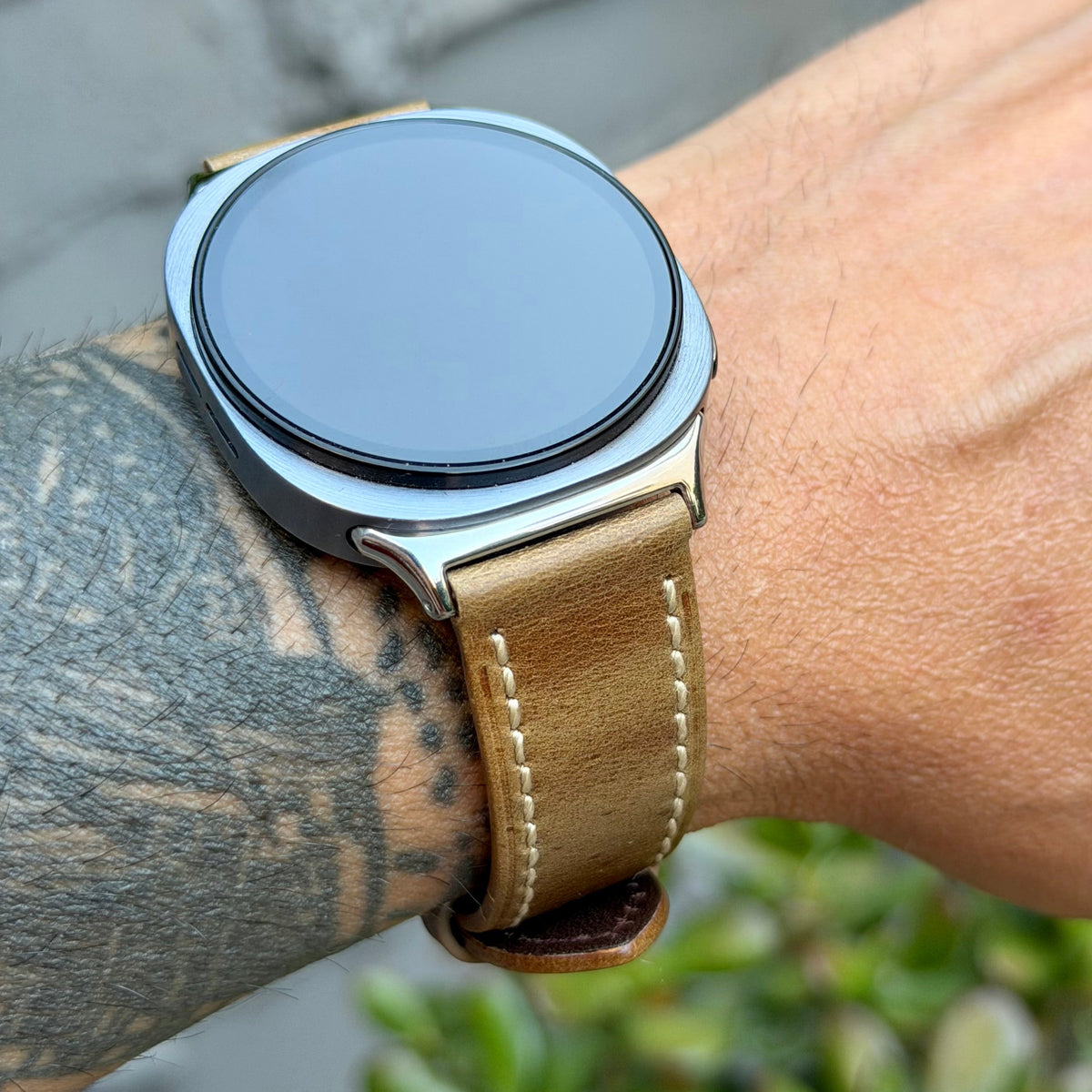 A close-up of a smartwatch with a round silver-toned case and a tan leather strap worn on a person's wrist. The background shows blurred outdoor greenery and gray pavement.