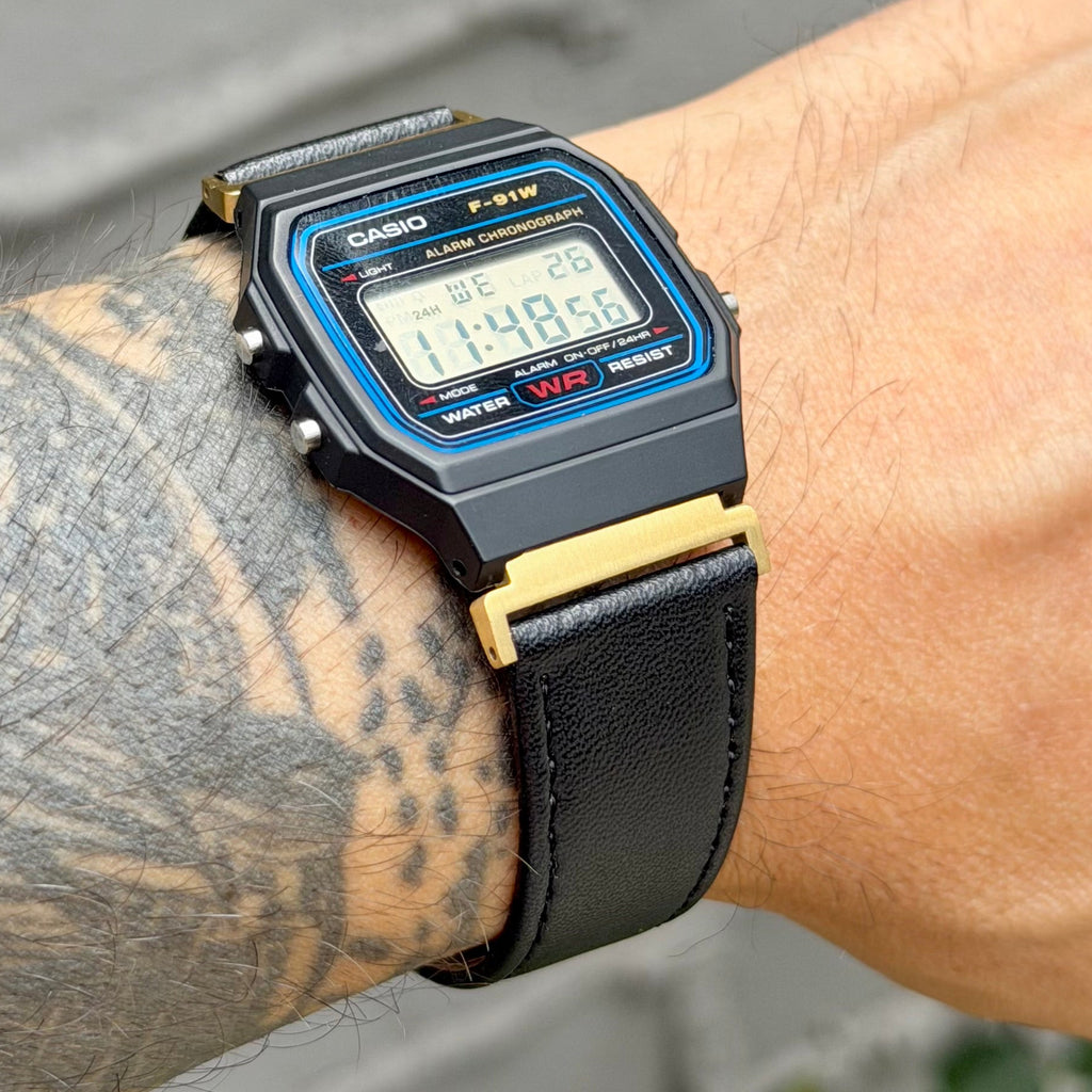 A close-up of a person's tattooed wrist wearing a black Casio F-91W digital watch with a black strap, blue bezel details, and gold-tone metal accents.