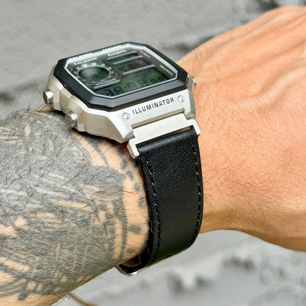 A close-up of a wrist wearing a rugged digital watch with a silver-toned metal case, black bezel, black leather strap, and a dark display. The word 'ILLUMINATOR' is visible on the side of the case.