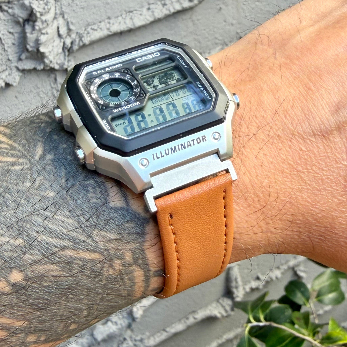 A wristwatch with a silver-tone metal case, black bezel, blue digital display, and tan leather strap worn on a tattooed arm against a gray concrete wall.