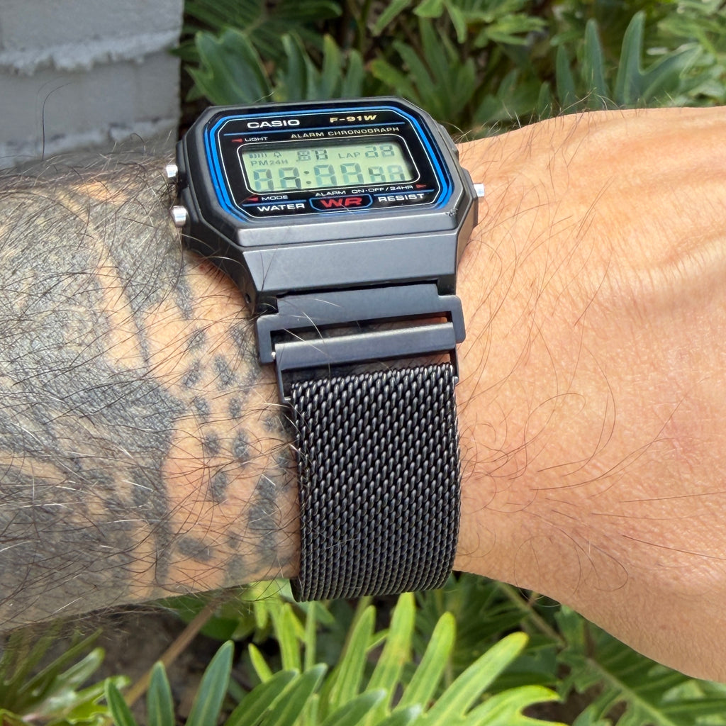 A black Casio F-91W digital wristwatch with a blue bezel and a black mesh strap, worn on a tattooed arm outdoors in front of green foliage.