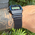 A black Casio F-91W digital wristwatch with a blue bezel and a black mesh strap, worn on a tattooed arm outdoors in front of green foliage.