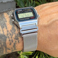 A silver Casio digital alarm watch with a mesh metal band worn on a person's wrist, with green foliage and a gray wall in the background.