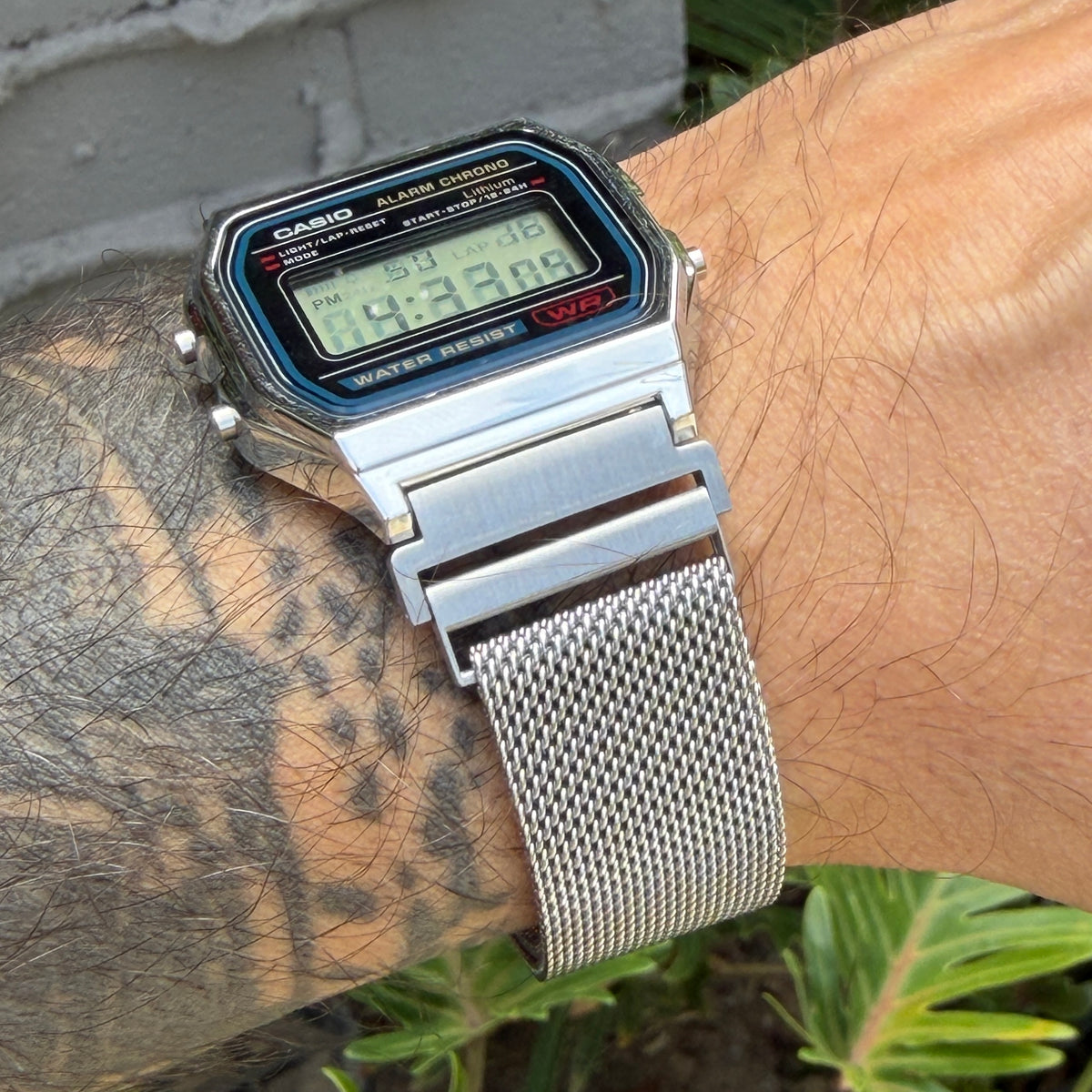 A silver Casio digital wristwatch with a mesh metal band worn on a tattooed wrist, photographed outdoors with a gray brick wall and green plants in the background.