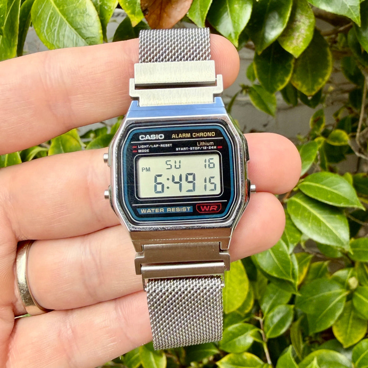 A vintage Casio digital alarm watch with a blue and silver metal case and mesh strap, held in a person's hand against a background of green leaves.