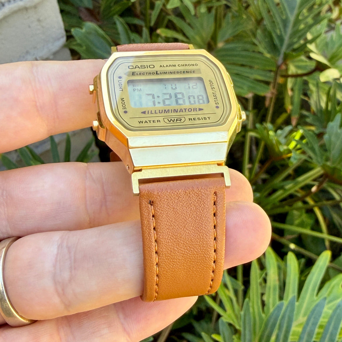 A gold-tone Casio digital watch with a brown leather strap, held in a person's hand against a background of green foliage.