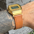 A gold-tone Casio digital watch with a tan leather strap worn on a person's wrist, photographed outdoors against a gray brick wall.