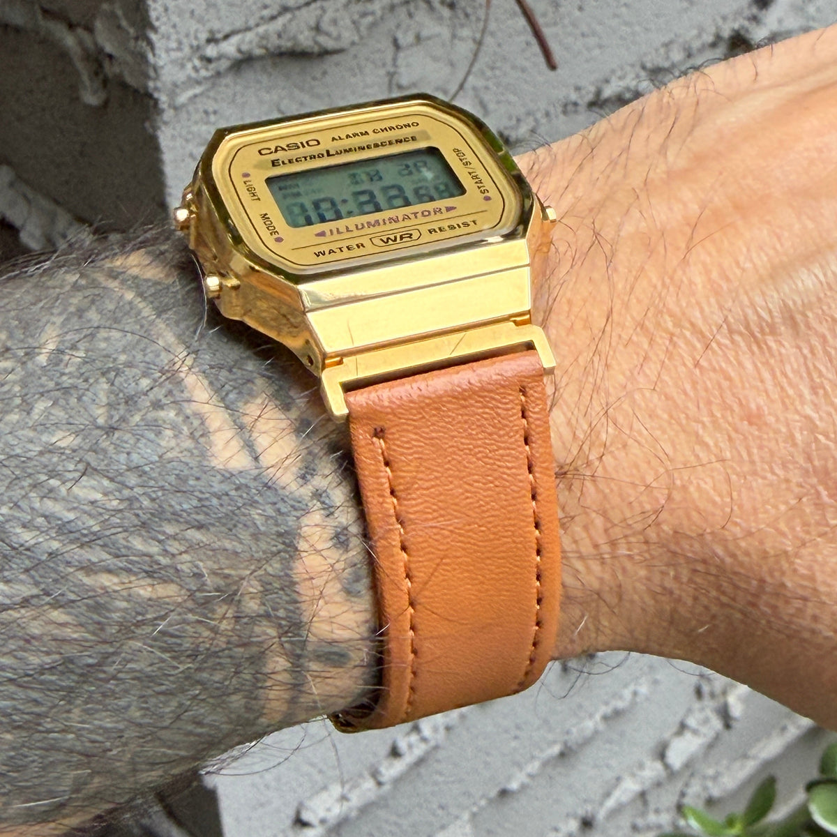 A gold-tone Casio digital watch with a tan leather strap worn on a person's wrist, photographed outdoors against a gray brick wall.