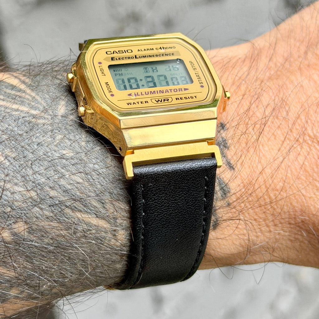 A gold-tone Casio digital watch with a black textured strap worn on a tattooed wrist against a blurred gray background.