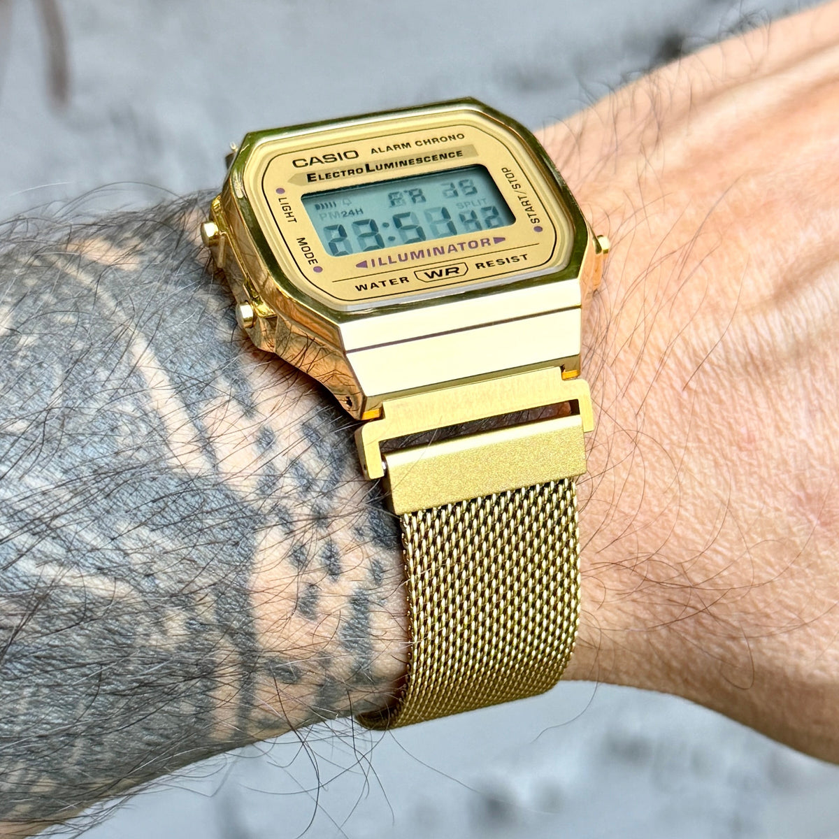 A gold-tone Casio digital watch with a gold mesh strap worn on a person's wrist, shown in a close-up outdoor shot.